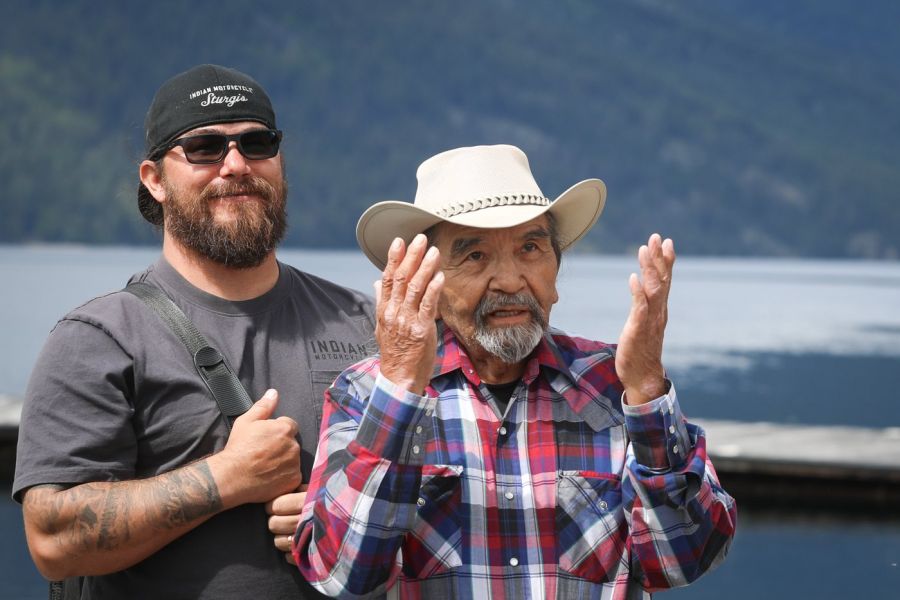 <who> Photo Credit: Aaron Hemens, Local Journalism Initiative Reporter</who>syilx Nation members caylx (Richard Armstrong), right, and his nephew nk’lxwcin (Chad Eneas) lead a ceremony as school children release juvenile salmon at takliʕaickst (Slocan Lake) on May 13. 